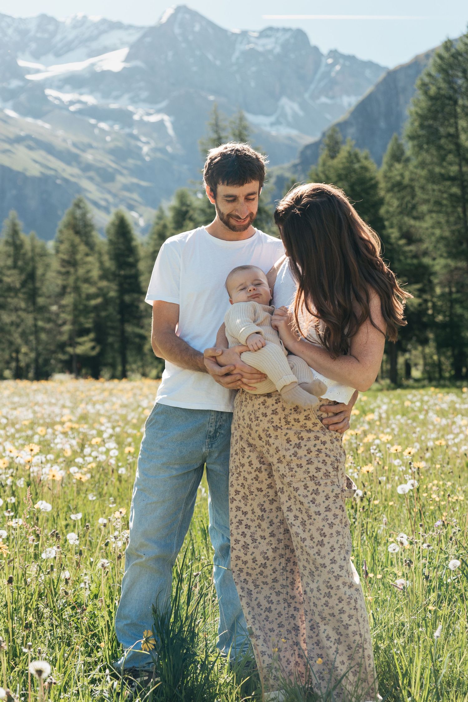 parents look at infant son in a field surrounded by French alps