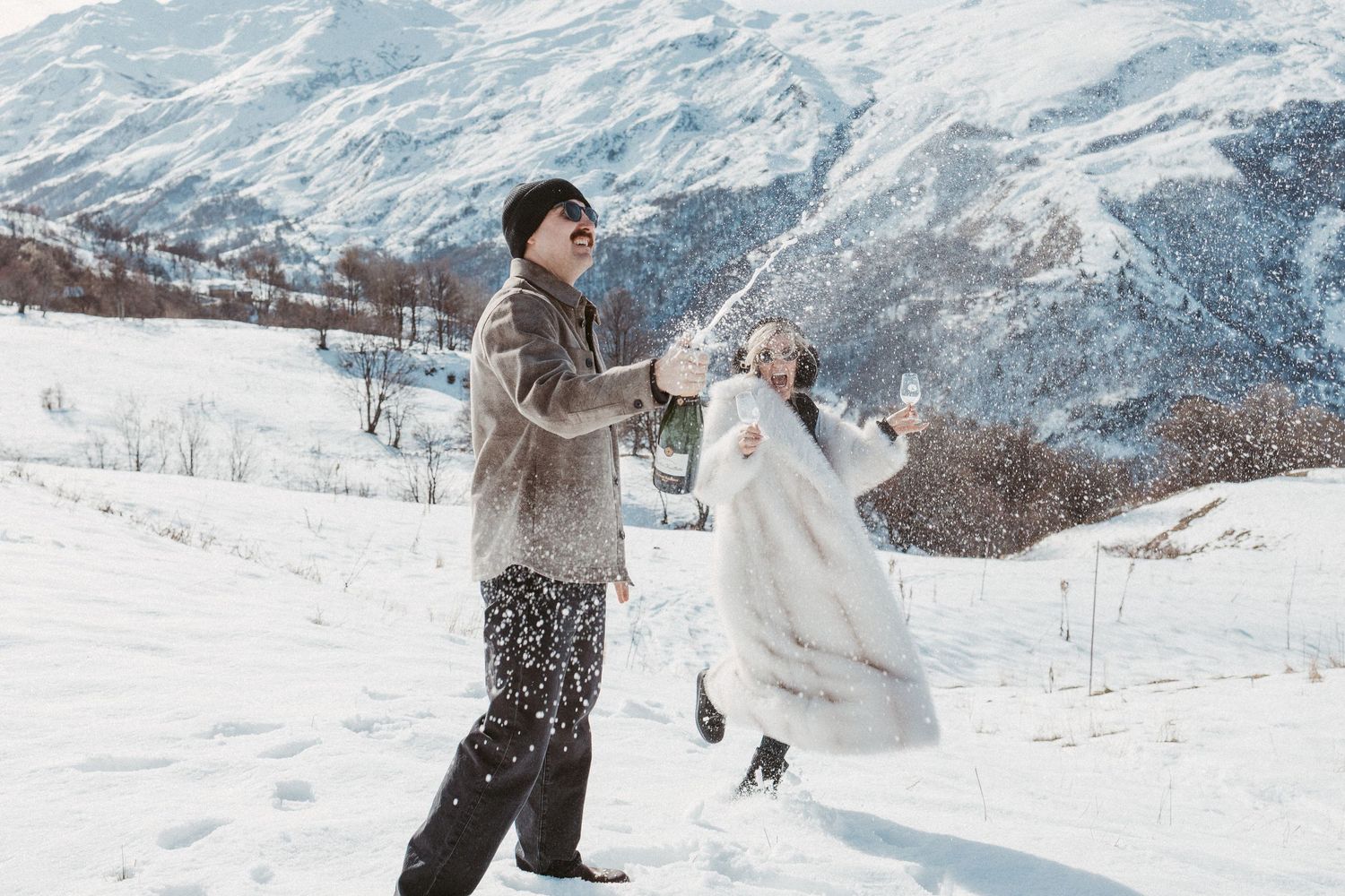 man pops champagne and sprays into air on top of mountain while fiancee cheers