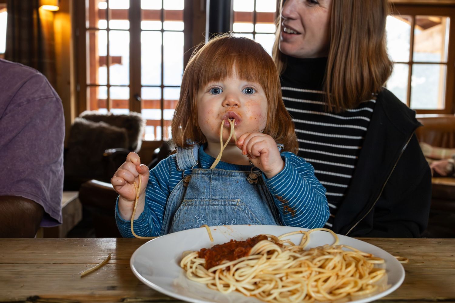 2 year old slurps spaghetti wearing blue dungarees