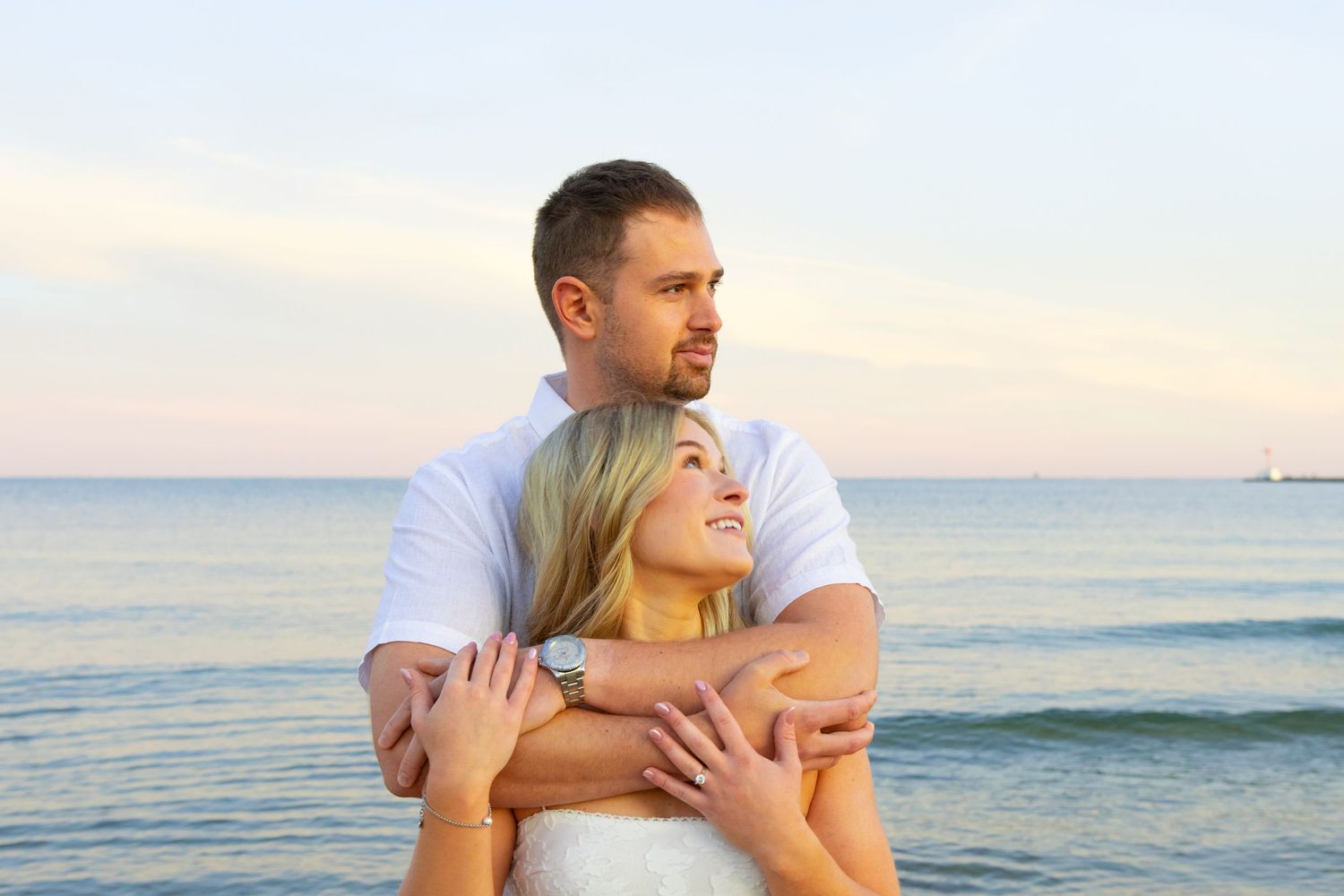 Romantic couple embracing on a beach at sunset, man in white shirt hugging woman from behind near the ocean.