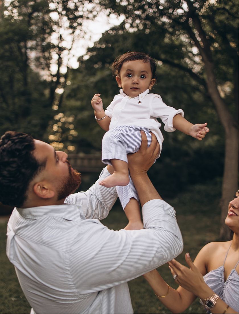 A joyful father lifts a smiling baby in the air outdoors while a woman watches, surrounded by lush green trees.