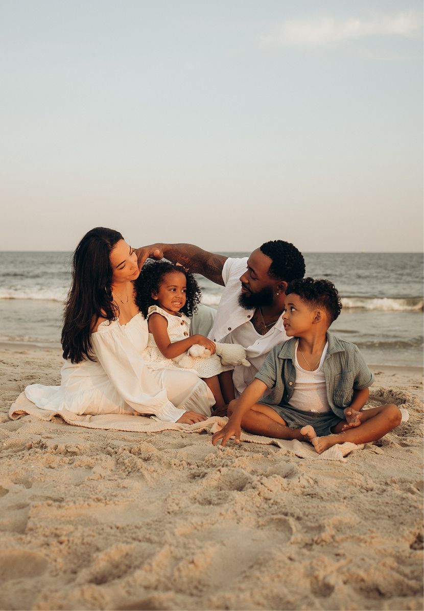 Family of four sitting together on sandy beach at sunset, parents and two young children in white and neutral outfits.