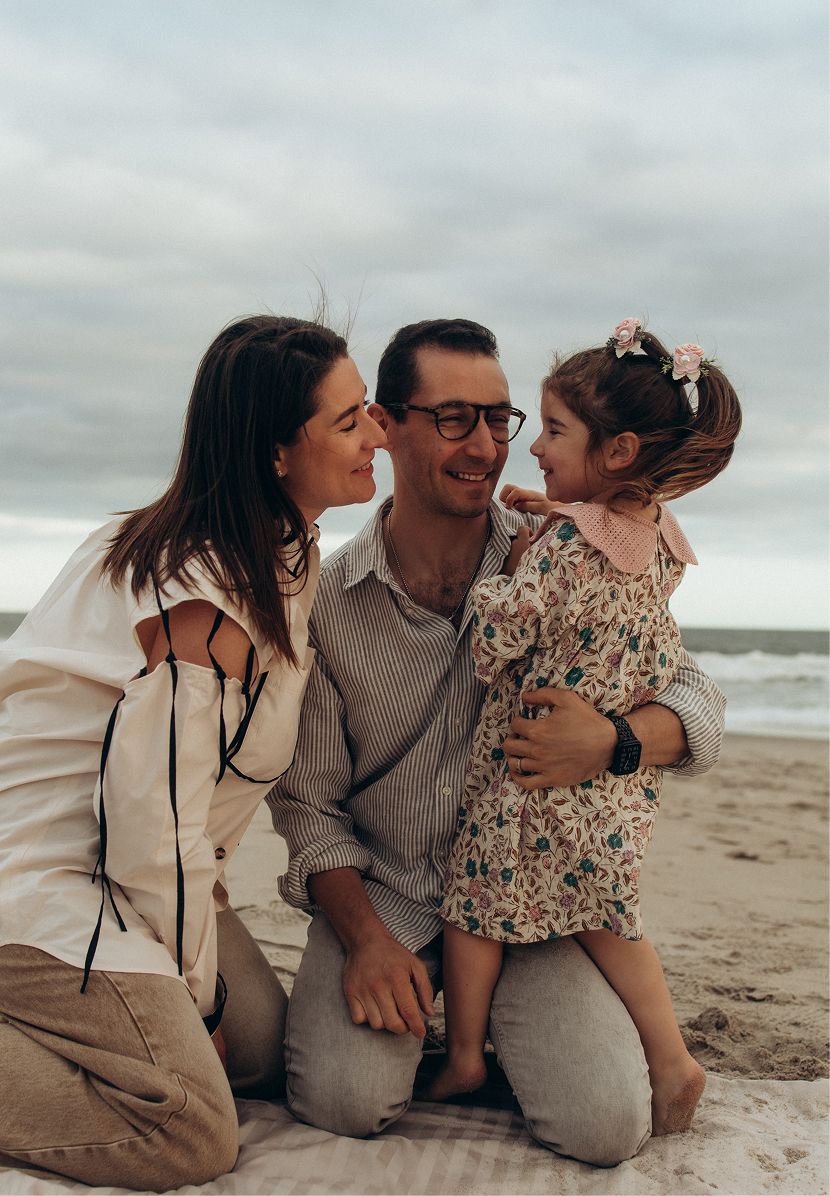 A happy family of three sits on a sandy beach, with parents smiling at their young daughter in a floral dress.