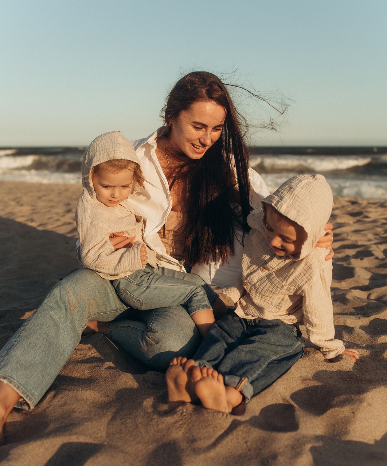 A smiling mother sits on a sandy beach with two young children in hooded jackets, enjoying a sunny day by the ocean.