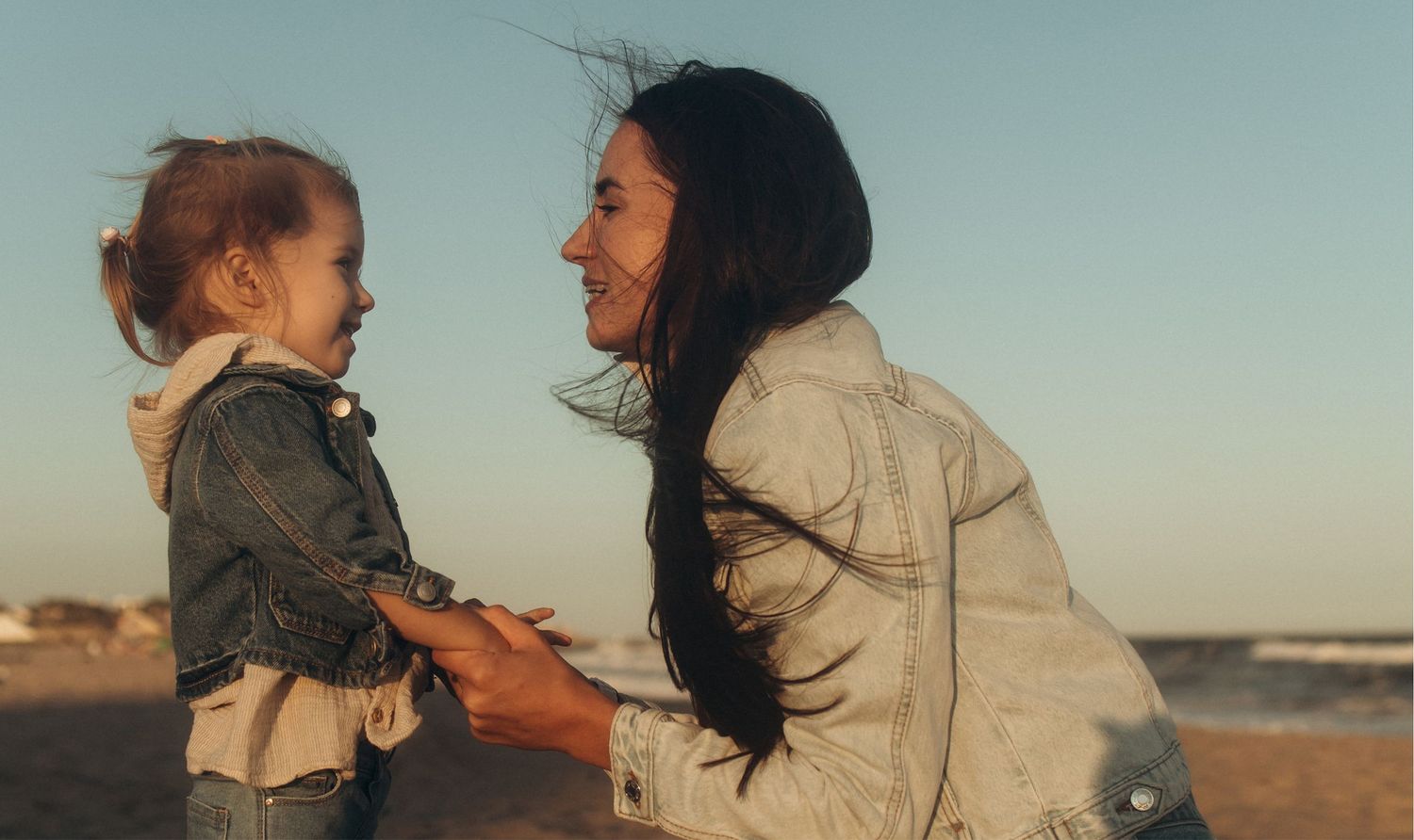 A smiling mother kneels to hold hands with her happy toddler daughter on a beach at golden hour.