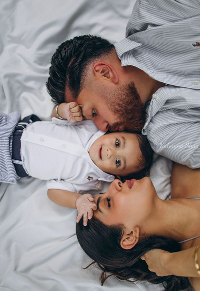 A couple lies on white bedding kissing their smiling baby between them in a tender family portrait.