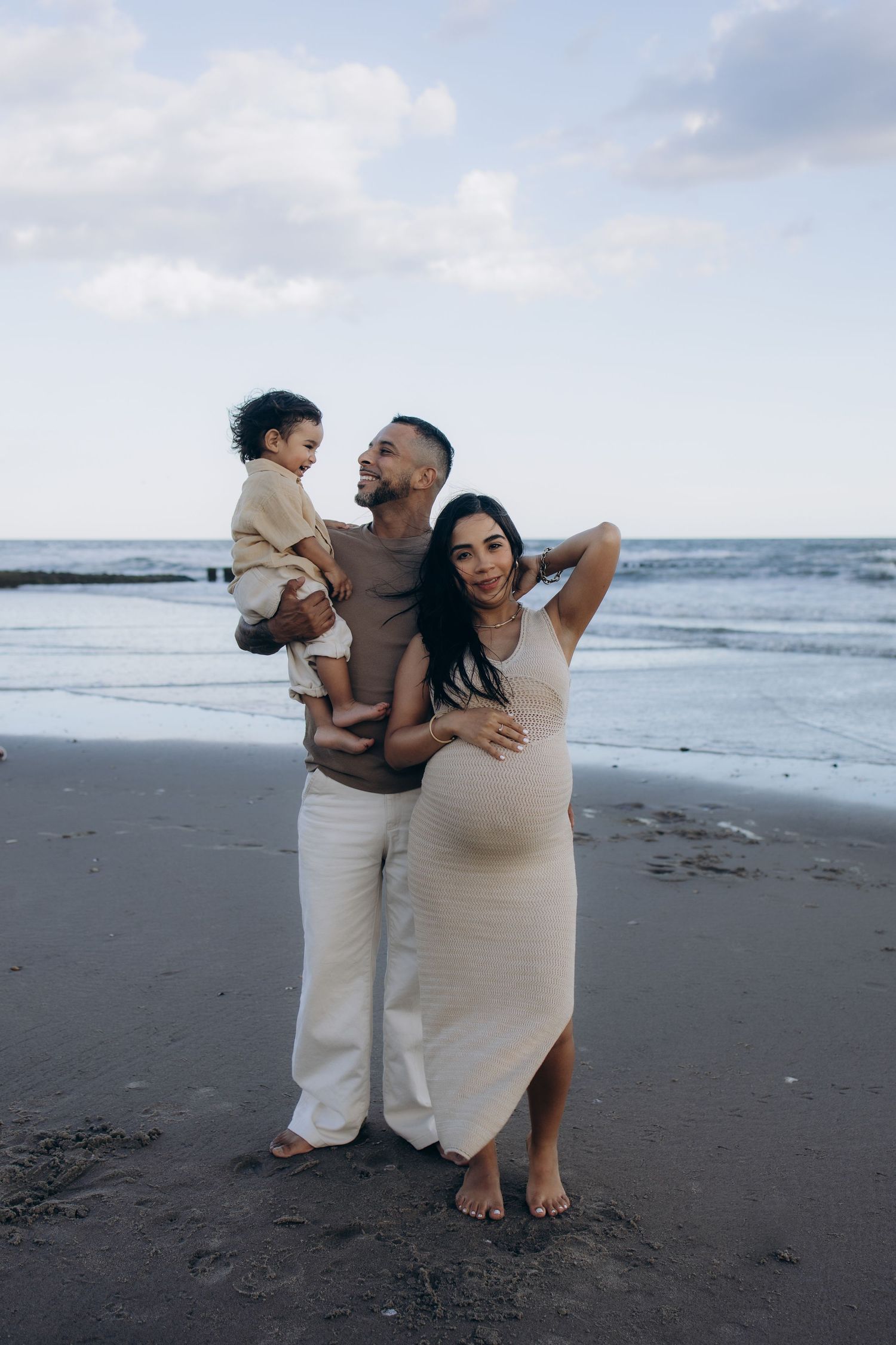 Pregnant woman with partner holding toddler posing together on a sandy beach with ocean waves in background.