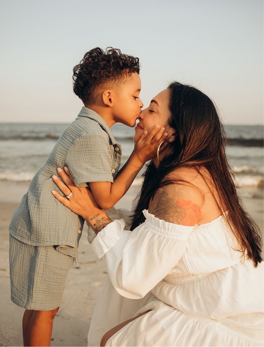 A mother and young son share a tender kiss on a sunny beach, she wears a white off-shoulder dress.