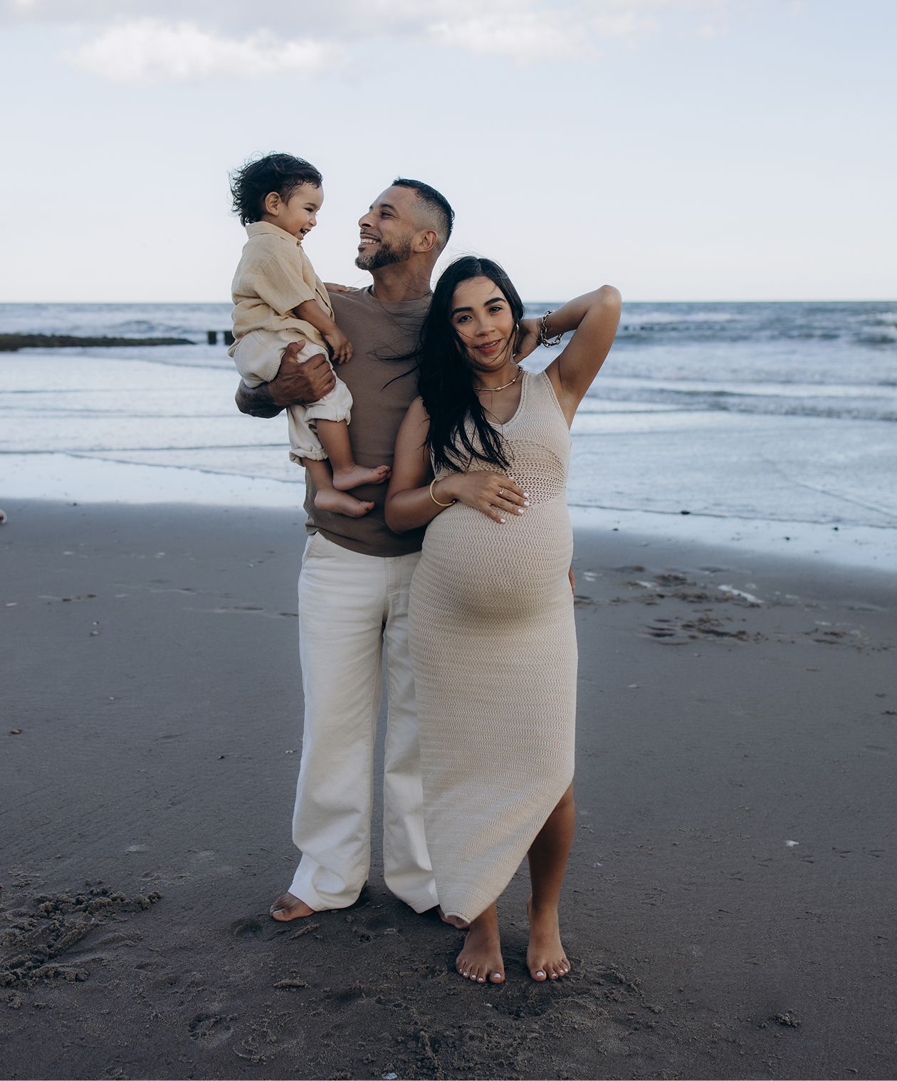 Pregnant woman in white dress poses with partner holding toddler on a sandy beach at dusk.