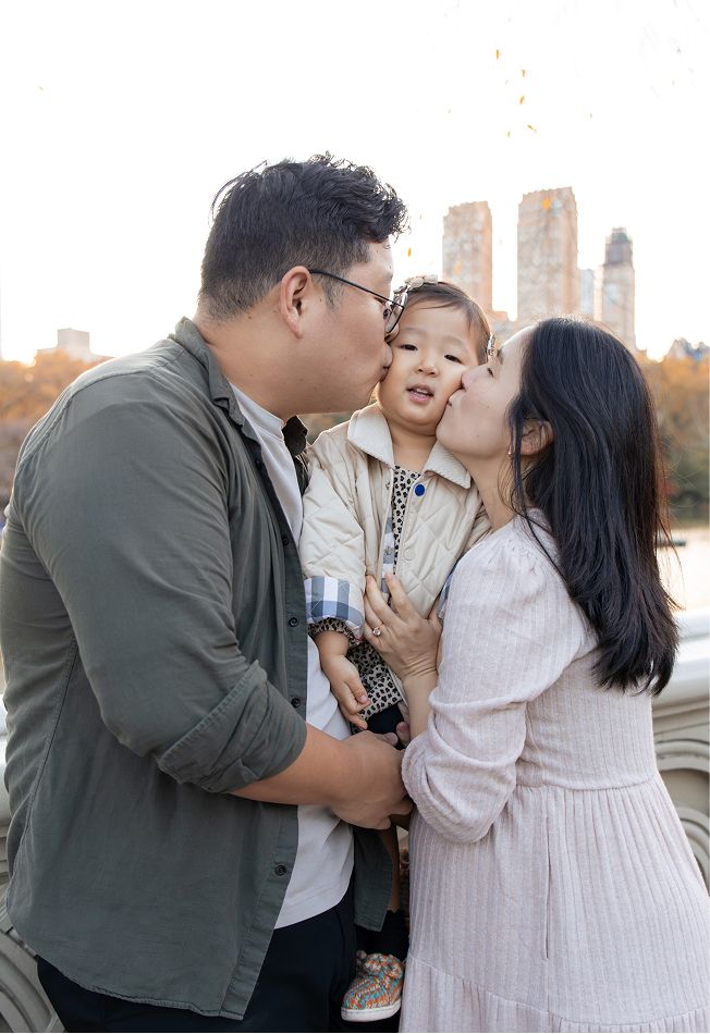 Parents kissing their toddler on the cheeks in Central Park with NYC skyline in the background.
