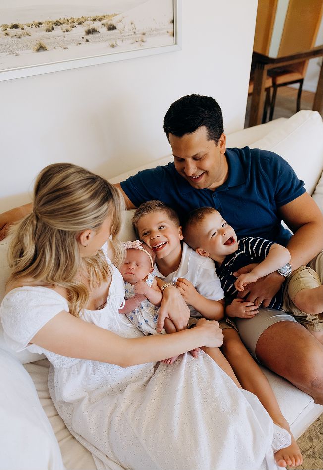 Happy family of five sitting on a white couch, parents smiling at their newborn baby held by two laughing young children.