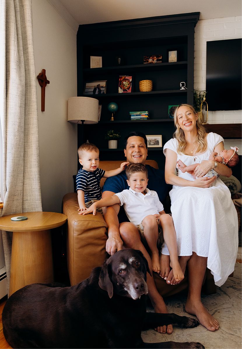 Happy family with two young boys, a baby, and a black Labrador posing in a cozy living room with dark bookshelves.