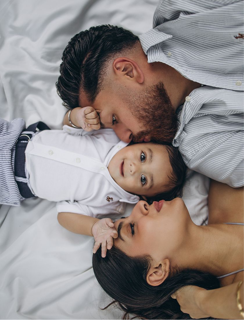 A loving family portrait with parents kissing their baby while lying on a white bed together.