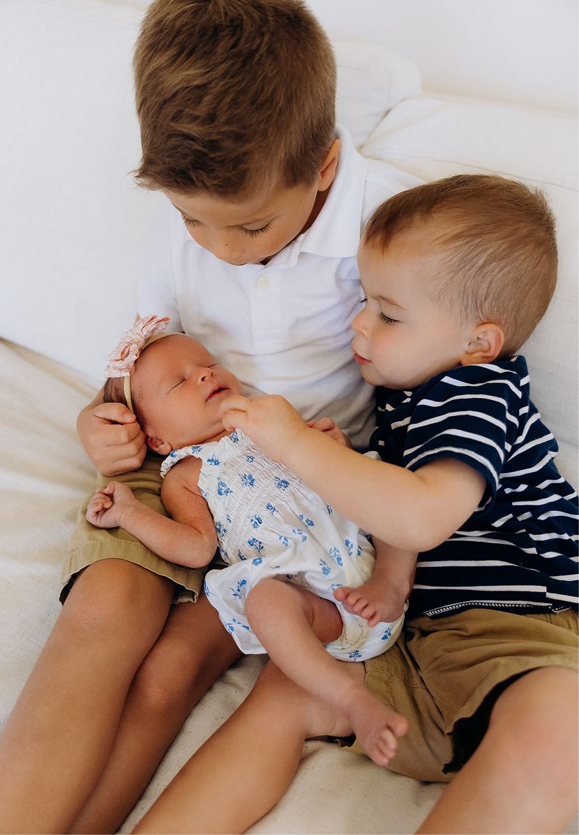 Two young boys tenderly hold their newborn sibling on a white bed in a heartwarming family portrait.