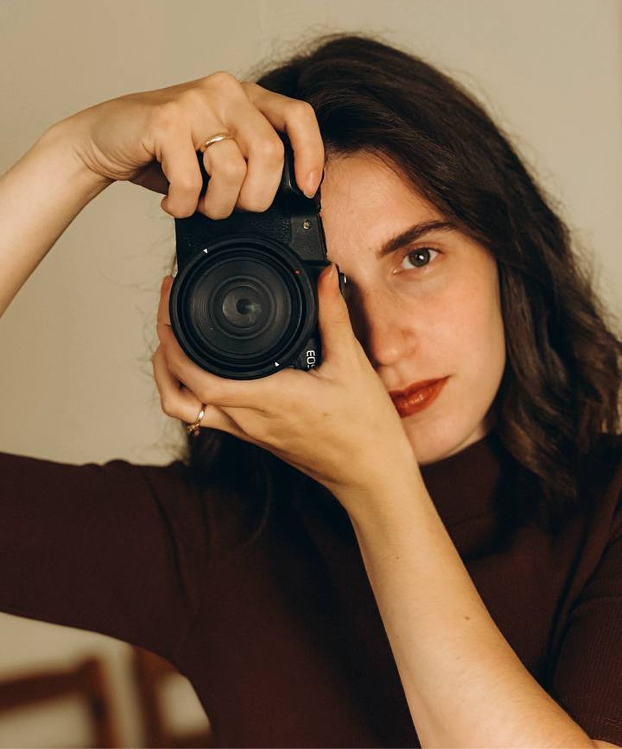 Young woman with dark hair holding a black camera up to her eye, wearing a brown turtleneck sweater.