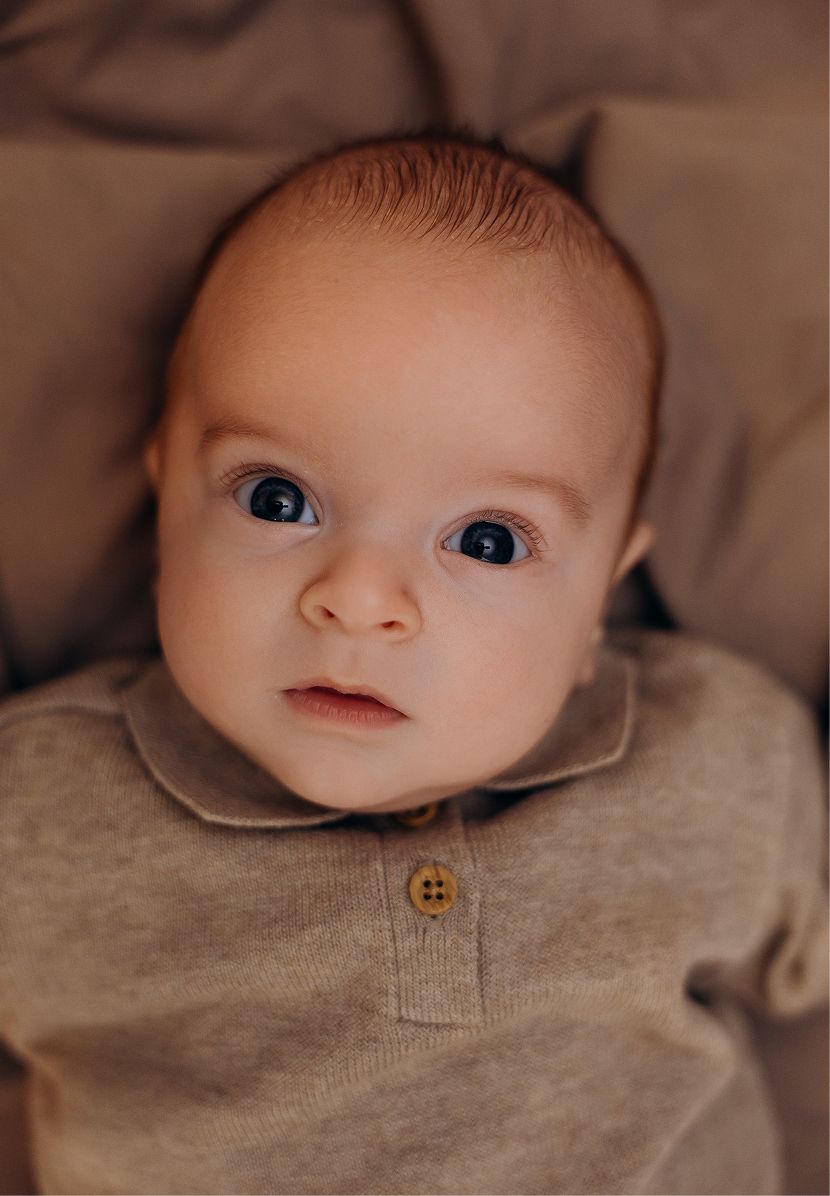 Adorable wide-eyed baby in a beige button collar onesie looking up with bright blue eyes.