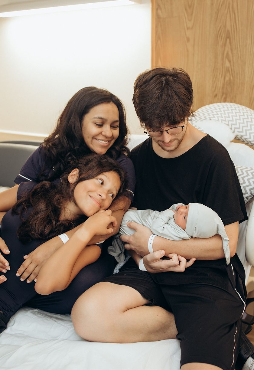 Family of four sitting on hospital bed, parents smiling at newborn baby wrapped in blanket with young child nearby.