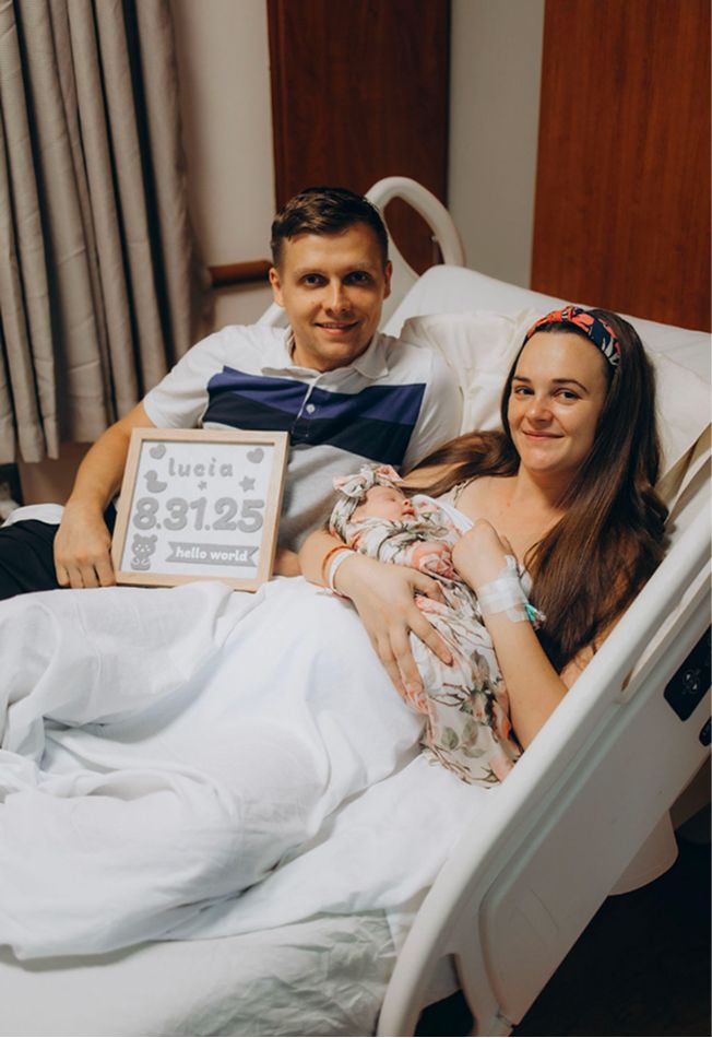 New parents in hospital bed holding newborn baby Lucia, born 8.31.25, smiling for a birth announcement photo.