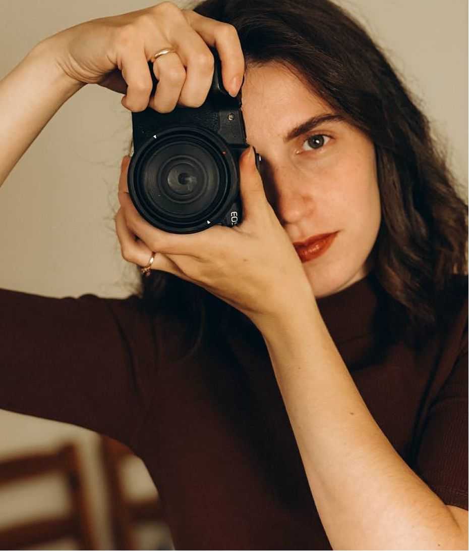 Woman holding a black camera up to her face, wearing a brown turtleneck, with dark wavy hair and a ring on her finger.