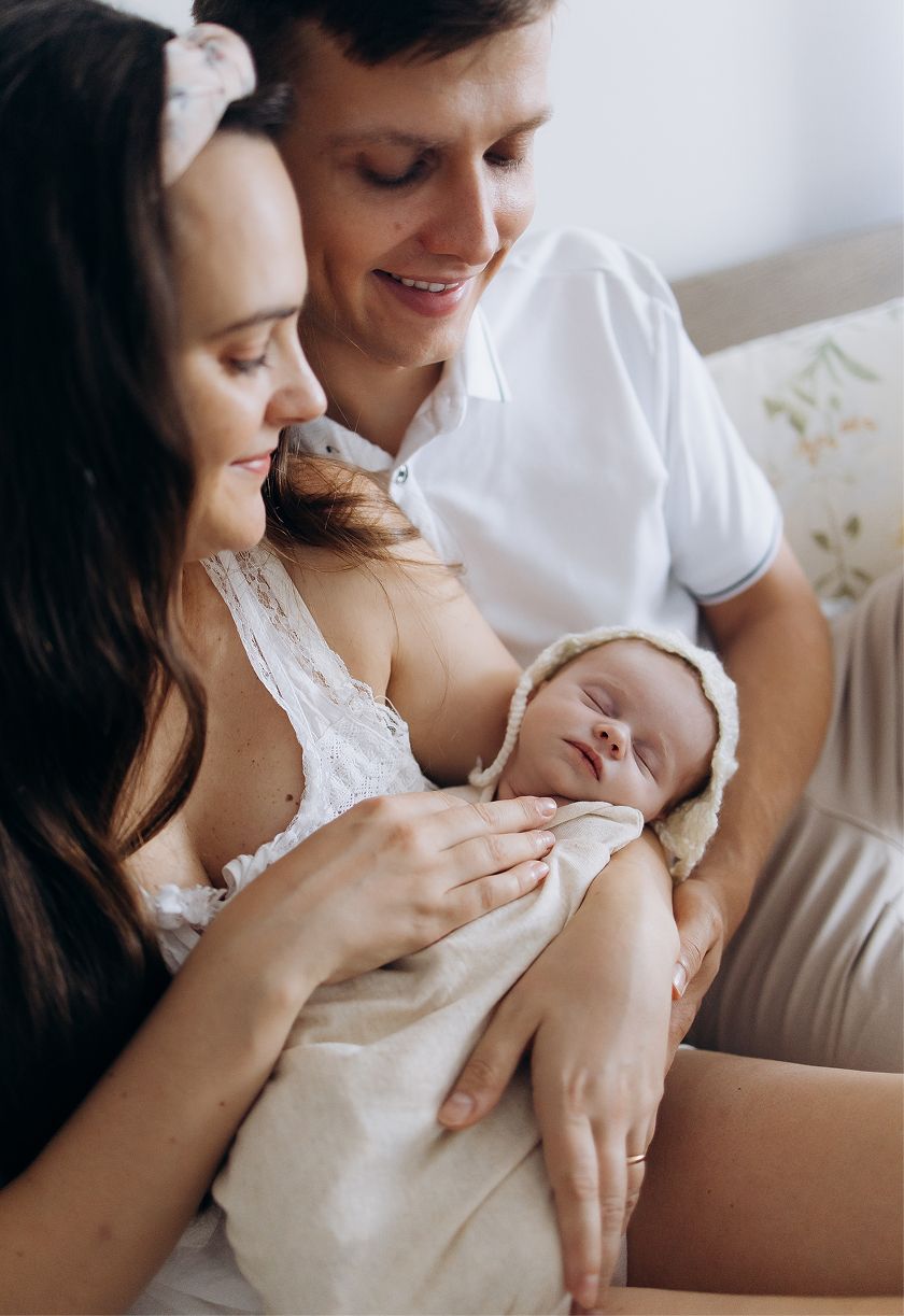 New parents smiling and gazing lovingly at their sleeping newborn baby wrapped in a white swaddle.