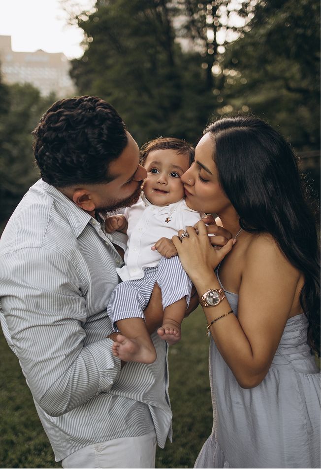 A couple kisses their smiling baby boy outdoors, with lush green trees in the background.