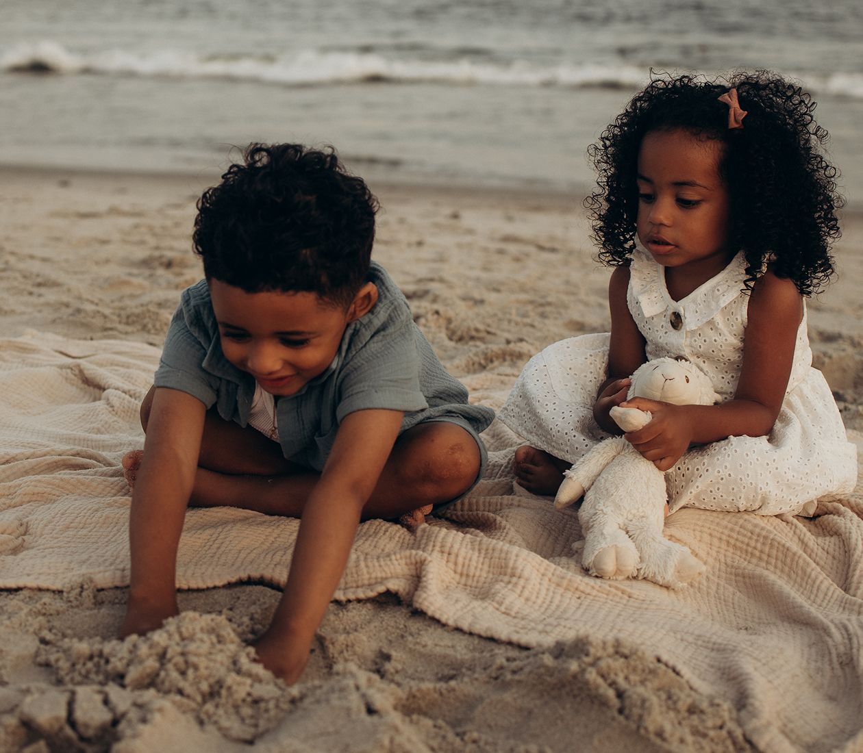 Two young children playing on a sandy beach, one digging in sand while the other holds a stuffed toy on a blanket.