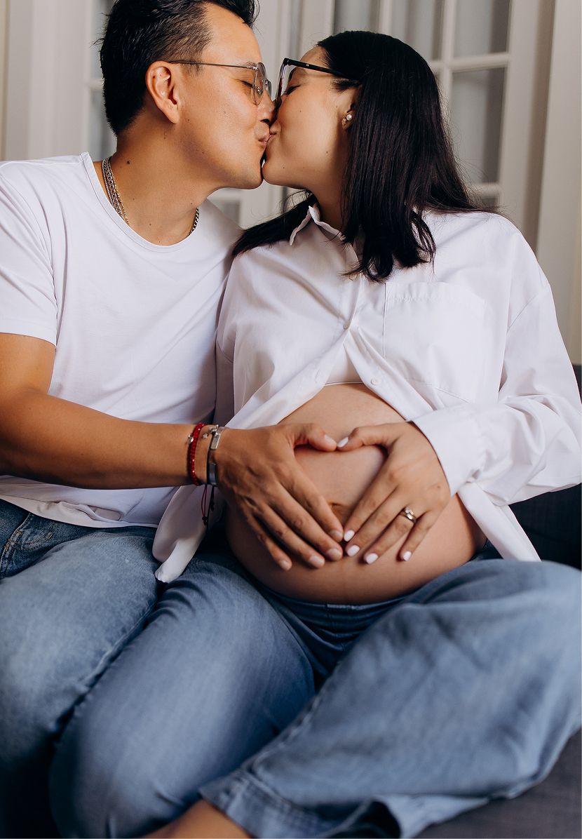 Couple sharing a kiss while pregnant woman forms heart shape on baby bump during maternity photo shoot.