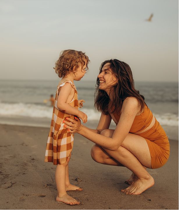 A woman in an orange bikini crouches on a beach, smiling at a toddler in a plaid outfit near ocean waves.