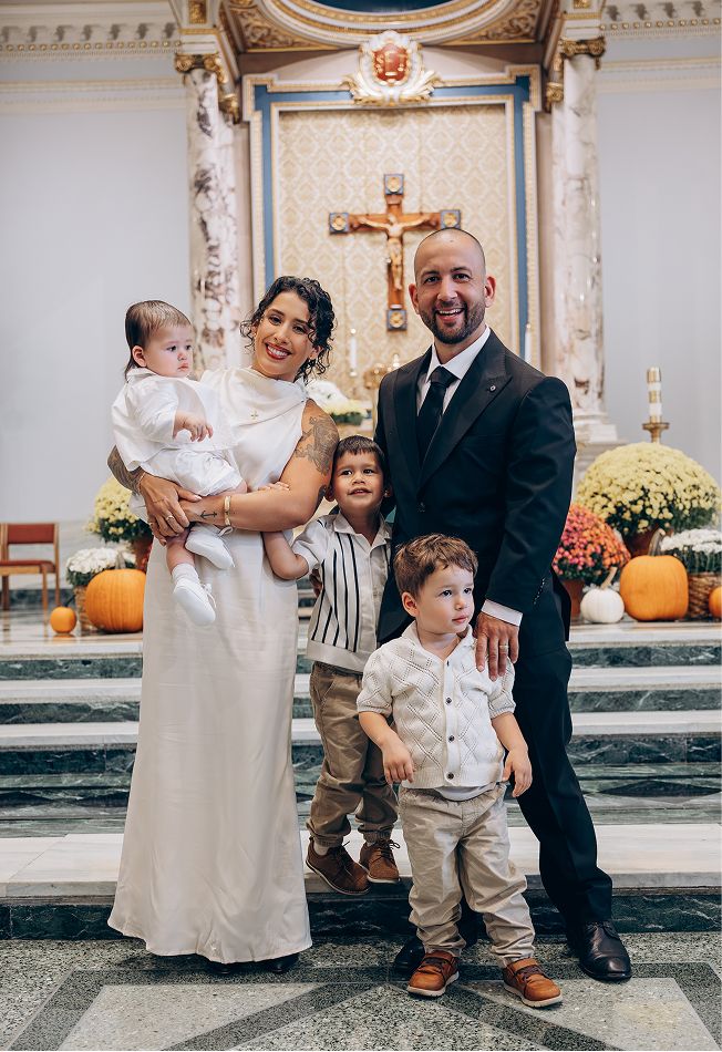 Family of five posing in a decorated Catholic church during a baptism ceremony, with a crucifix and fall flowers in the background.