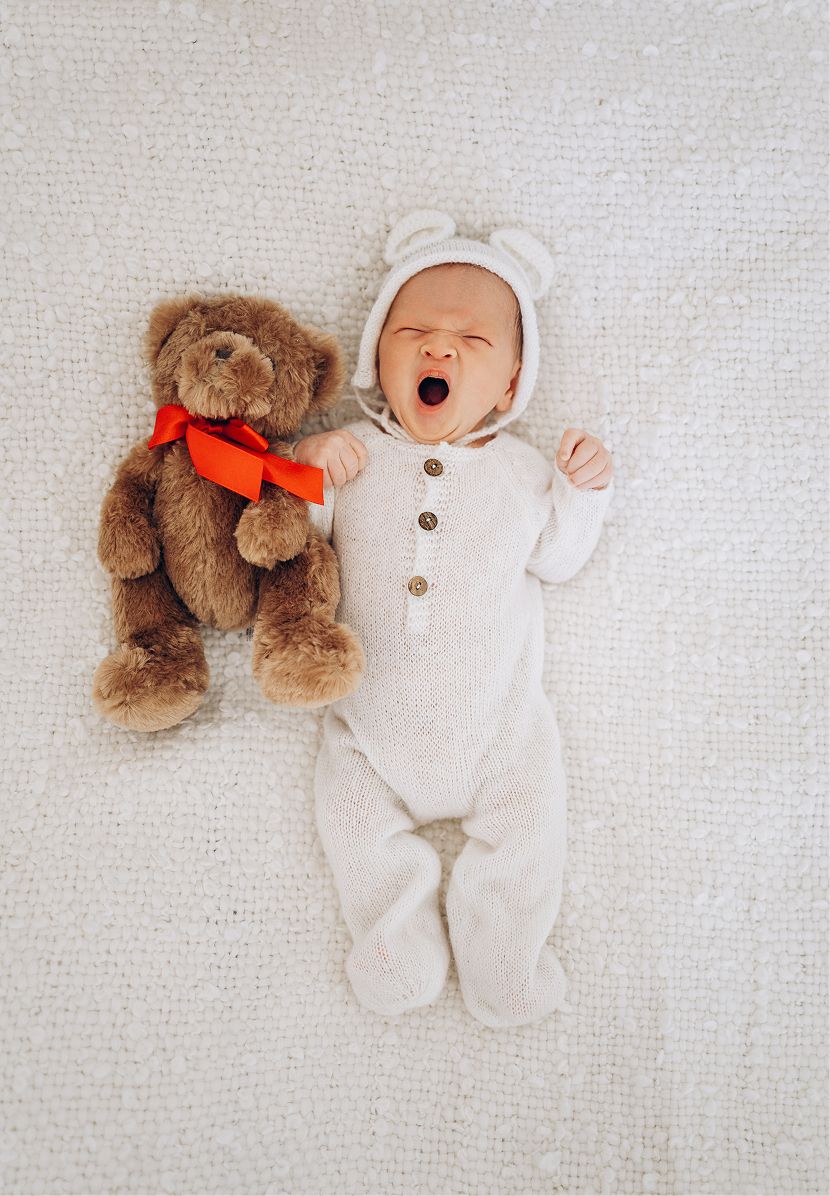 Yawning newborn baby in white bear onesie lying next to a brown teddy bear with red ribbon on white blanket.