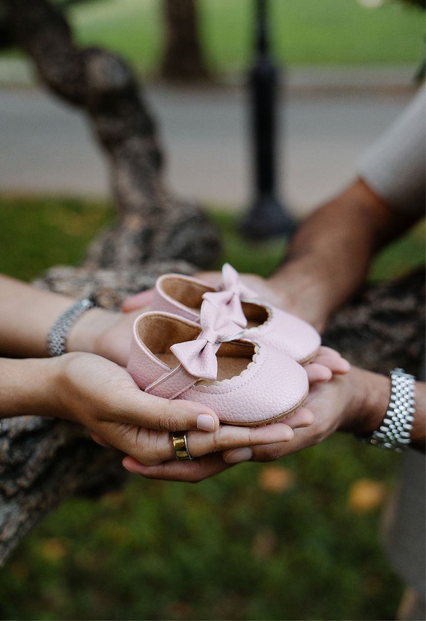 Two hands cradling tiny pink baby girl shoes with a bow, announcing a pregnancy outdoors near a tree.