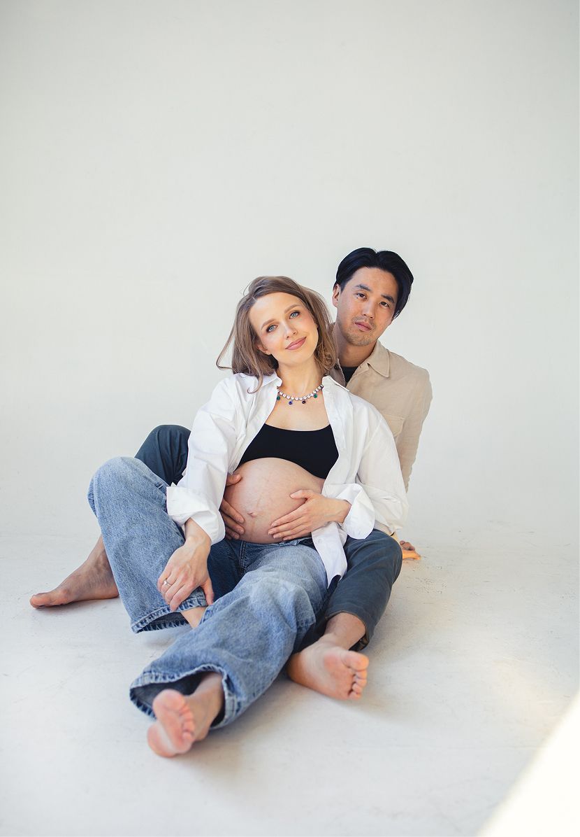Pregnant couple sitting on floor, woman in jeans and black crop top with open white shirt, man behind her.