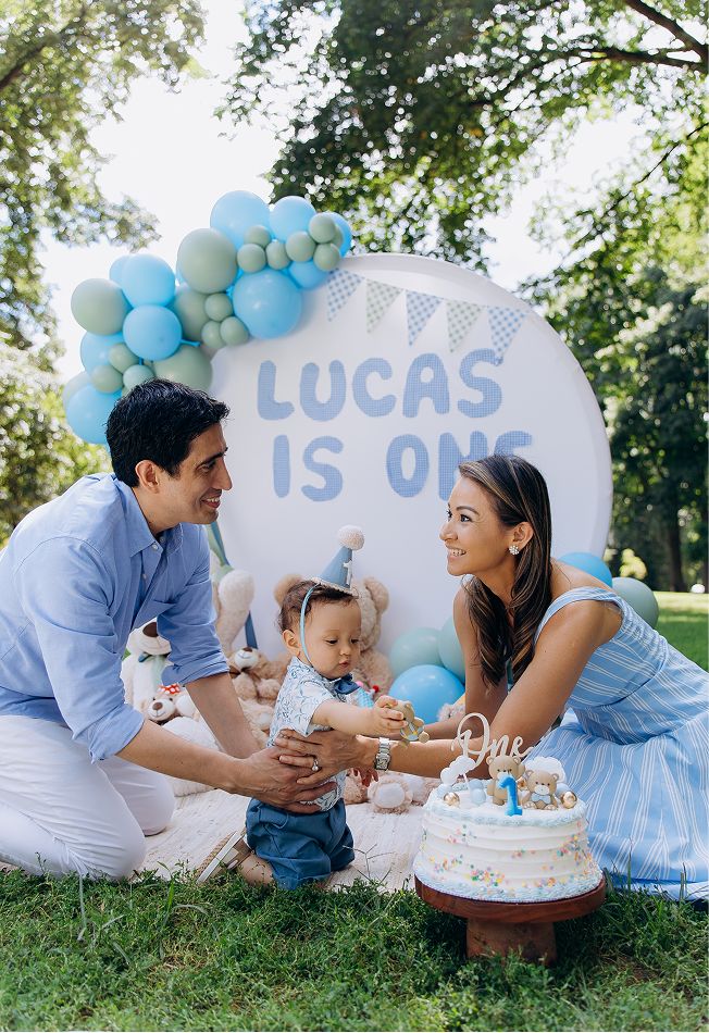A family celebrates baby Lucas turning one with a blue balloon arch, cake, and decorations outdoors.