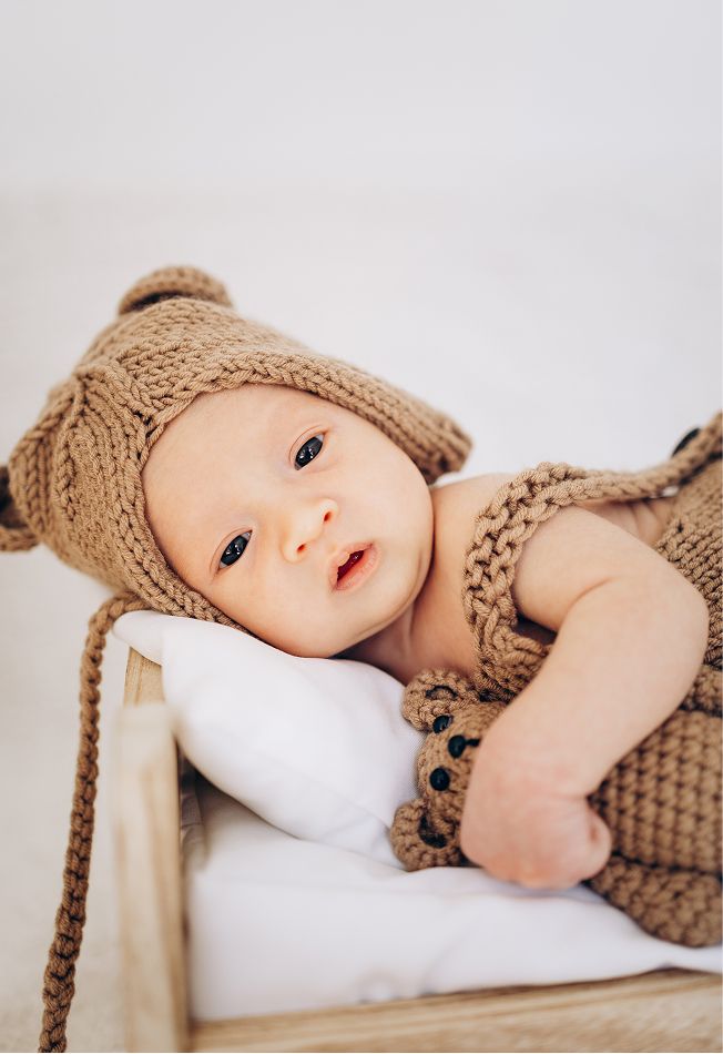 Adorable newborn baby wearing a knitted brown teddy bear outfit and hat, lying in a tiny wooden bed.