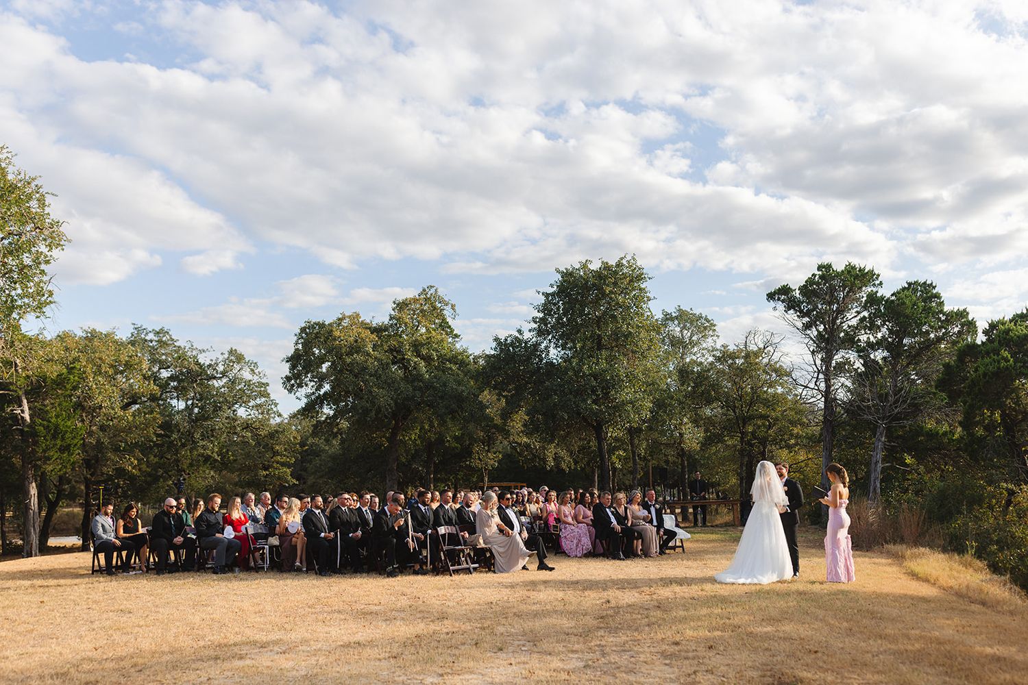 Outdoor wedding ceremony on a hilltop with guests seated under trees and cloudy sky in the background.