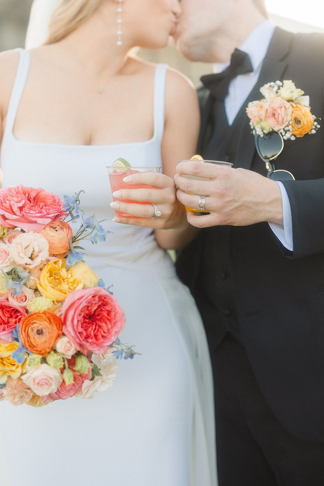 Newlywed couple holding drinks with vibrant coral and peach floral bouquet featuring garden roses and ranunculus.