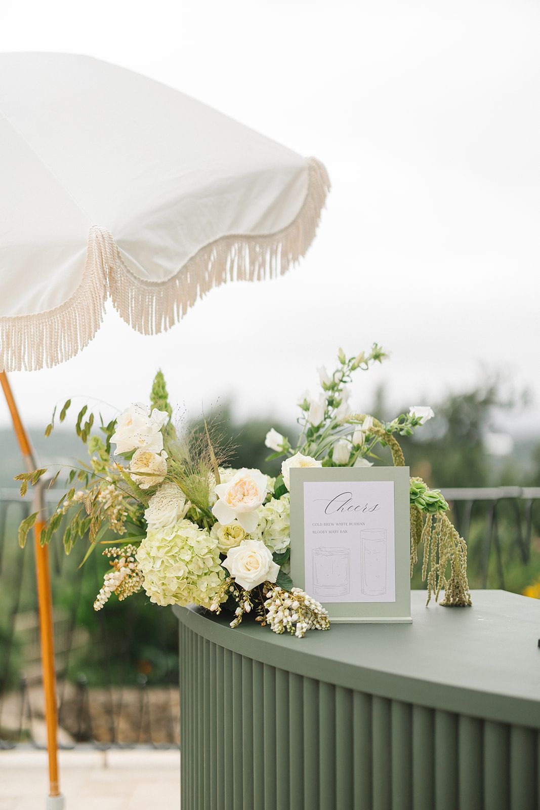 White parasols and floral arrangements featuring hydrangeas and roses adorn a sage green railing at an outdoor event.