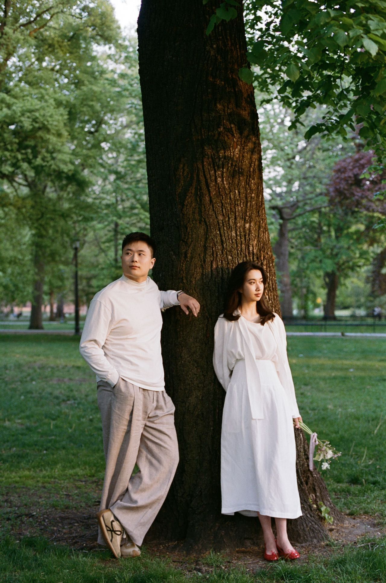 A couple poses by a large tree in a park, elegantly dressed in white. The man leans casually against the tree while the 