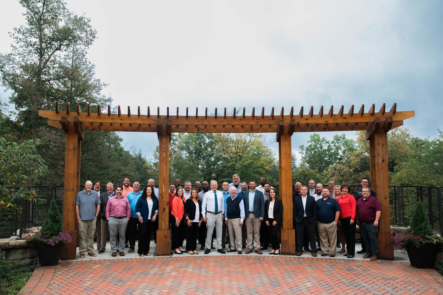 Large group of professionals posing under a wooden pergola in an outdoor brick patio setting.