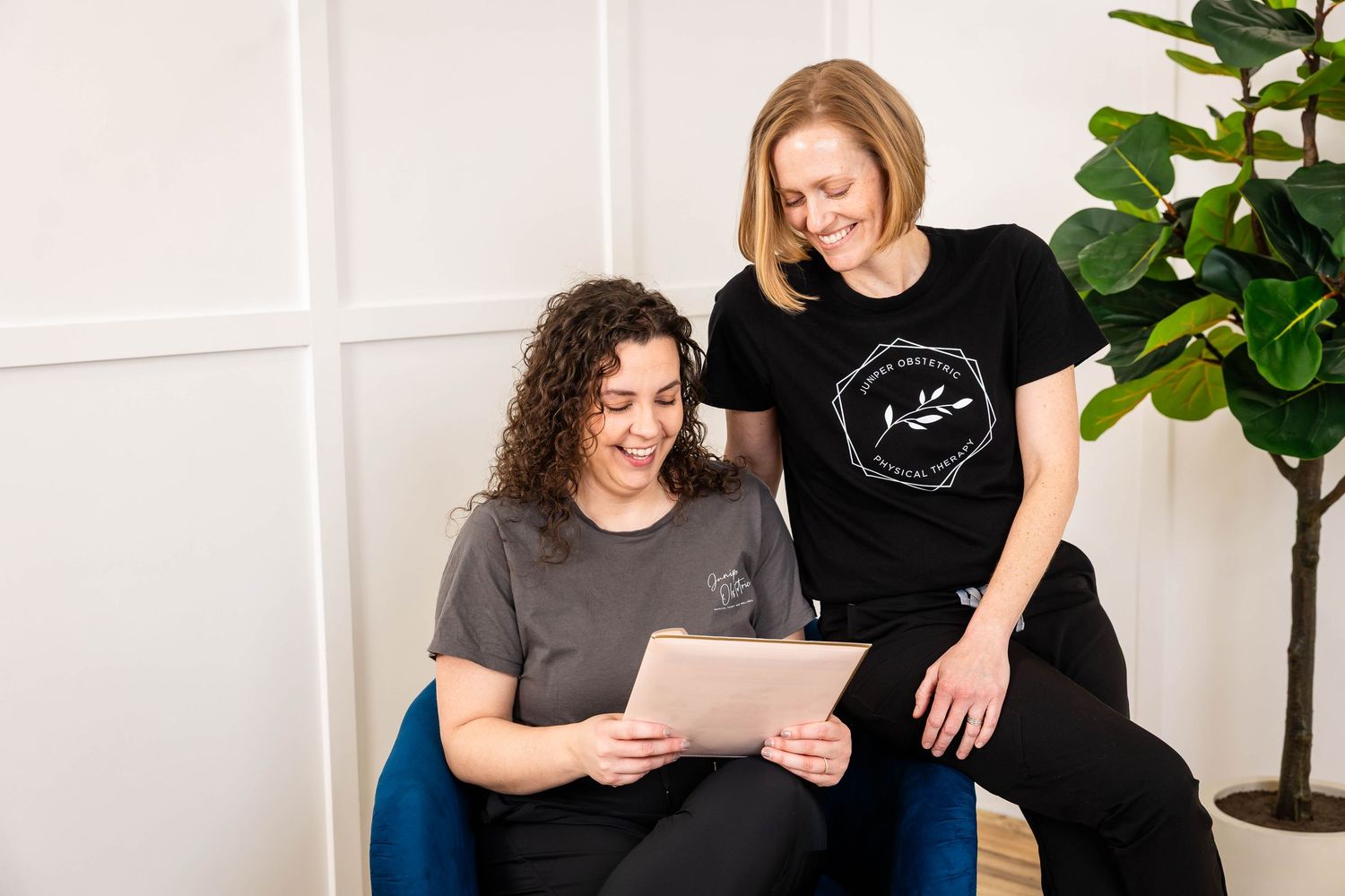 Two women in matching black branded t-shirts smile while reviewing content on a tablet in a bright modern office.