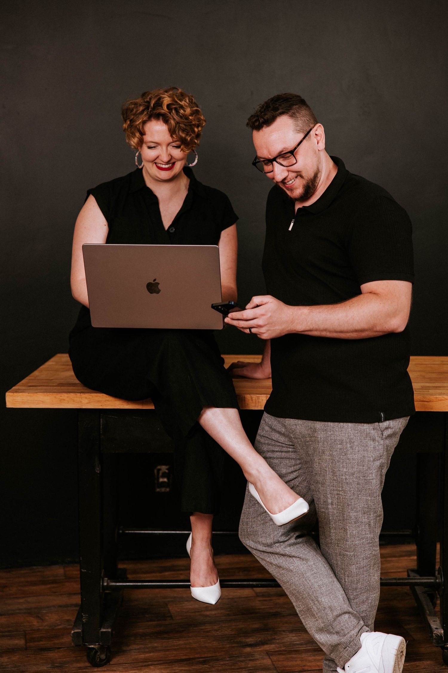 A woman with a laptop and a man with a smartphone sit on a wooden table, smiling and collaborating in a modern office.