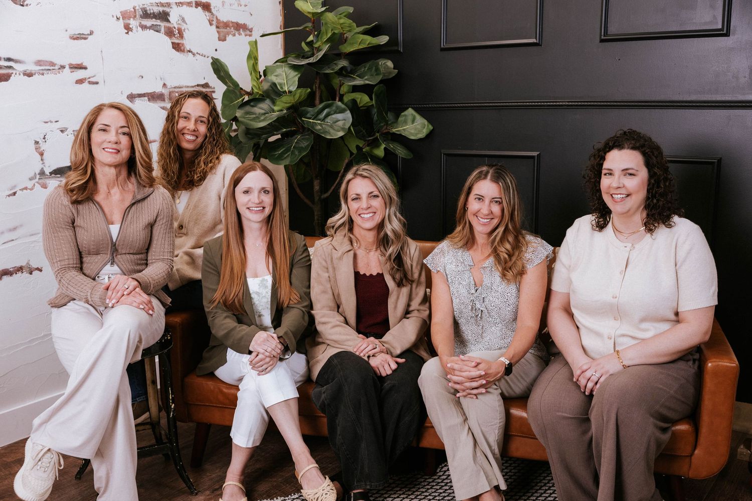 Six professional women smiling and posing together on a bench in a modern, stylish office setting.
