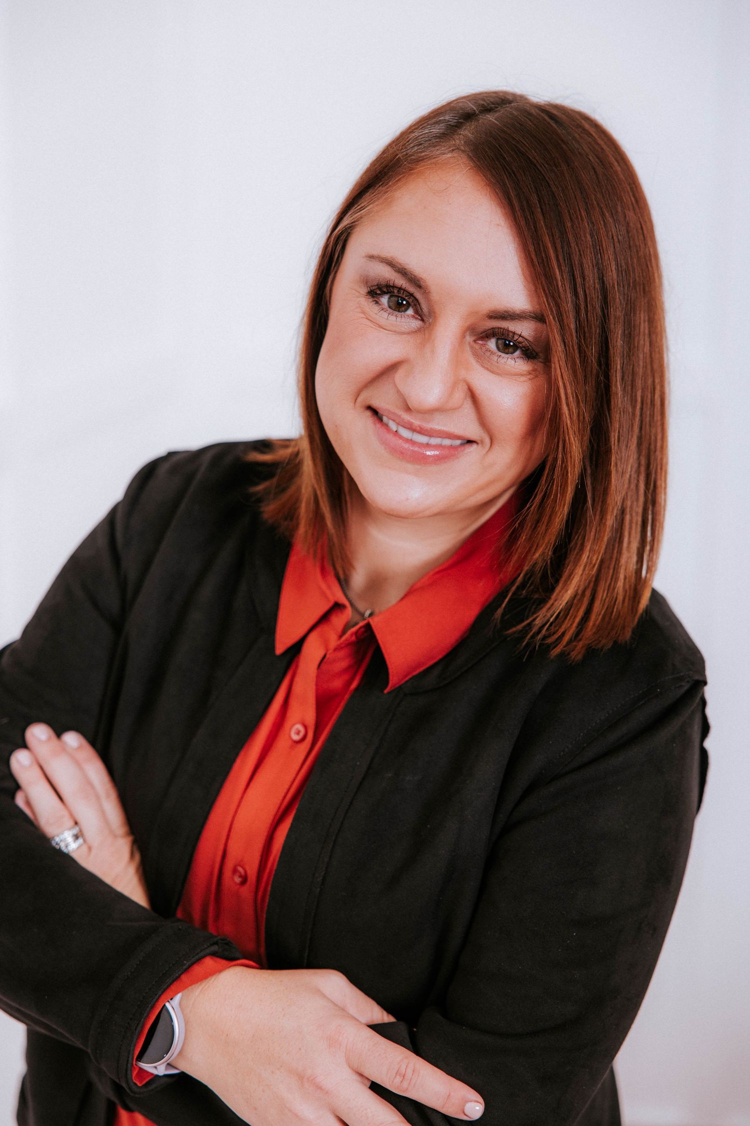 Professional woman in black blazer and red shirt smiling with arms crossed against white background.