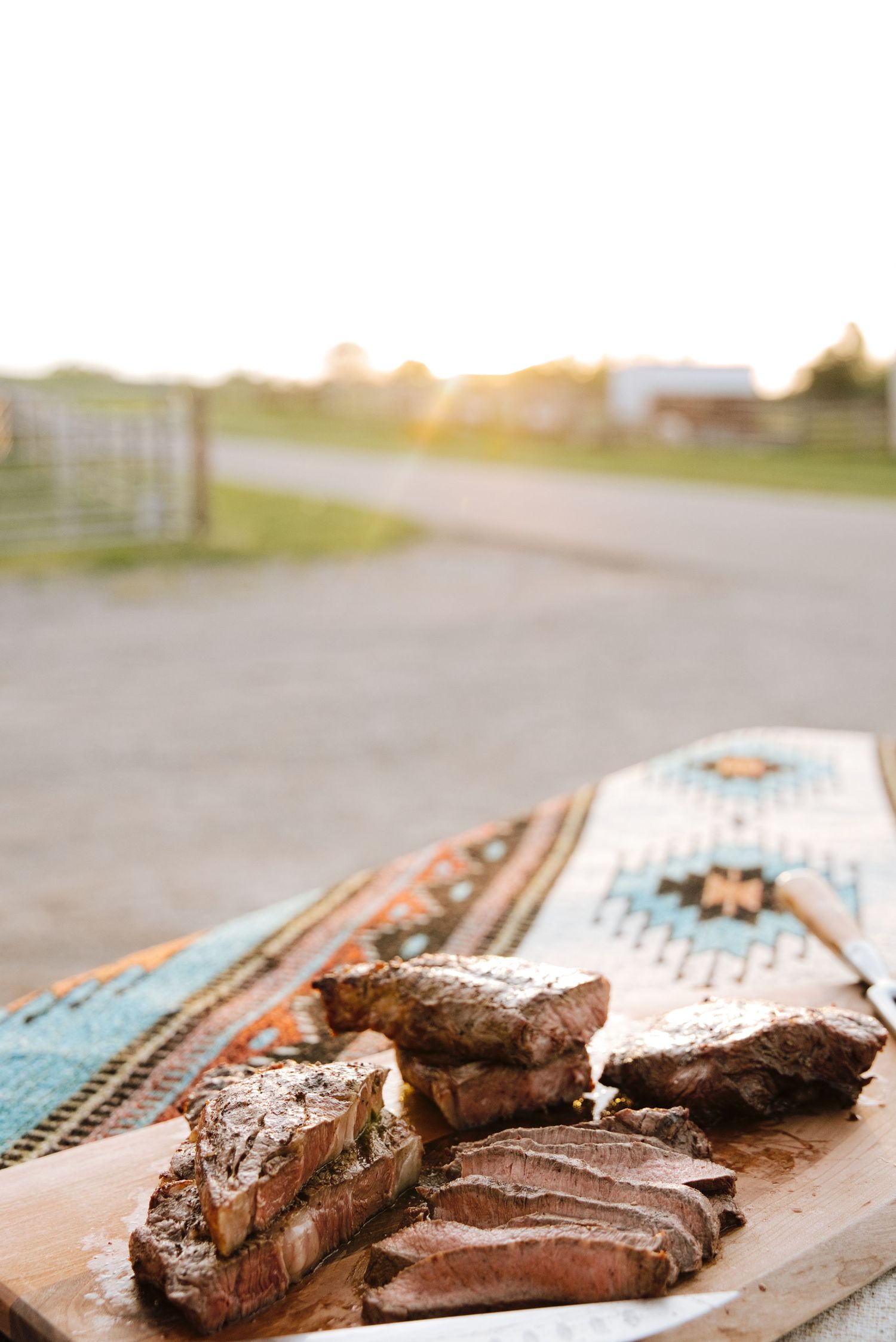 Sliced grilled steak on a wooden board with a colorful southwestern blanket at golden hour on a ranch.