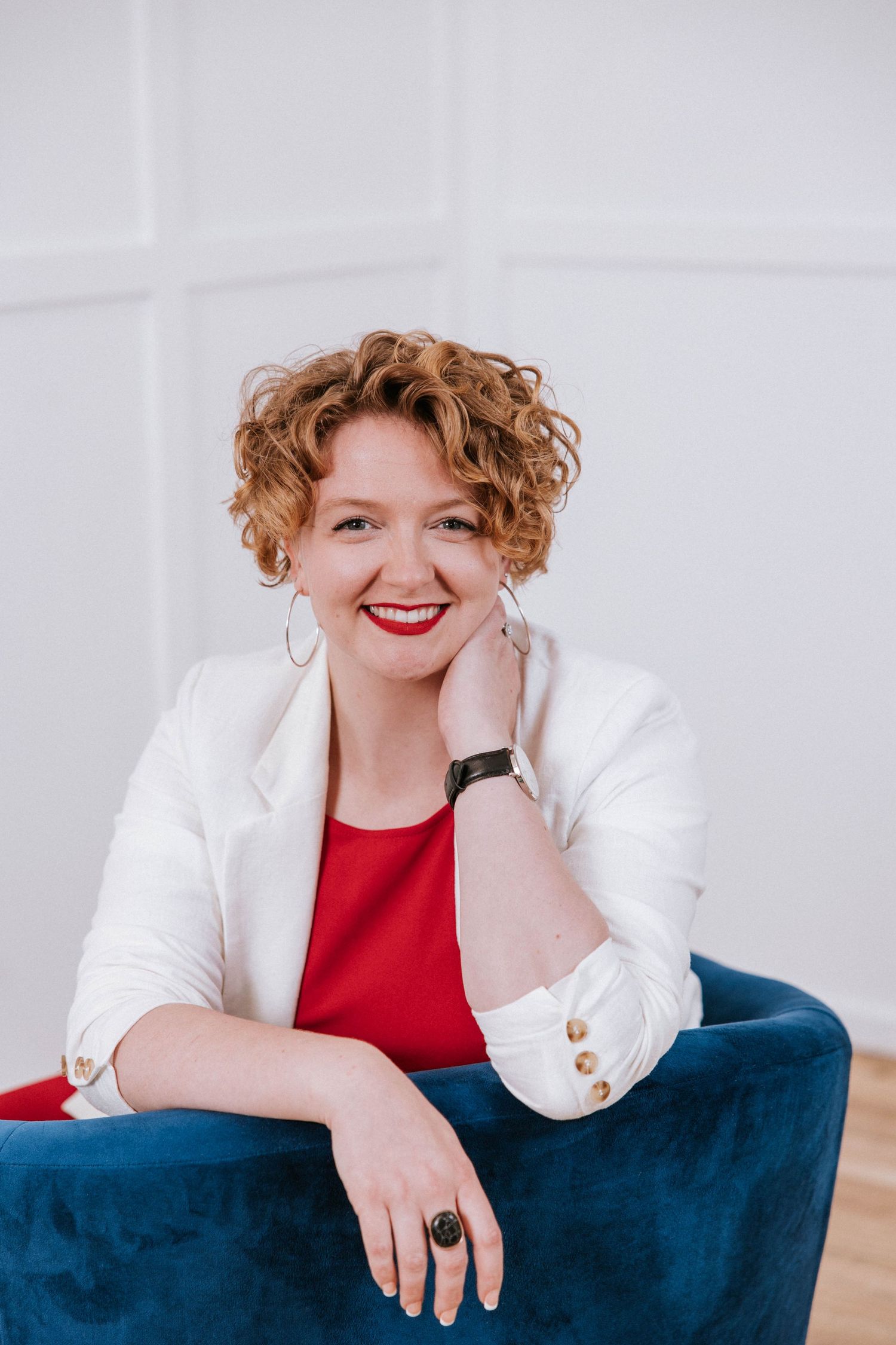 Smiling woman with curly red hair in white blazer and red top seated in blue velvet chair against white wall.