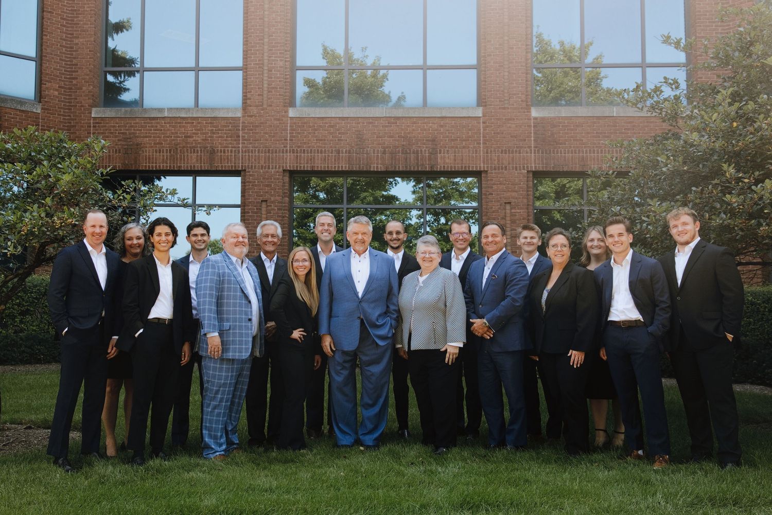 Professional corporate team posing outdoors in front of a brick office building, dressed in formal business attire.
