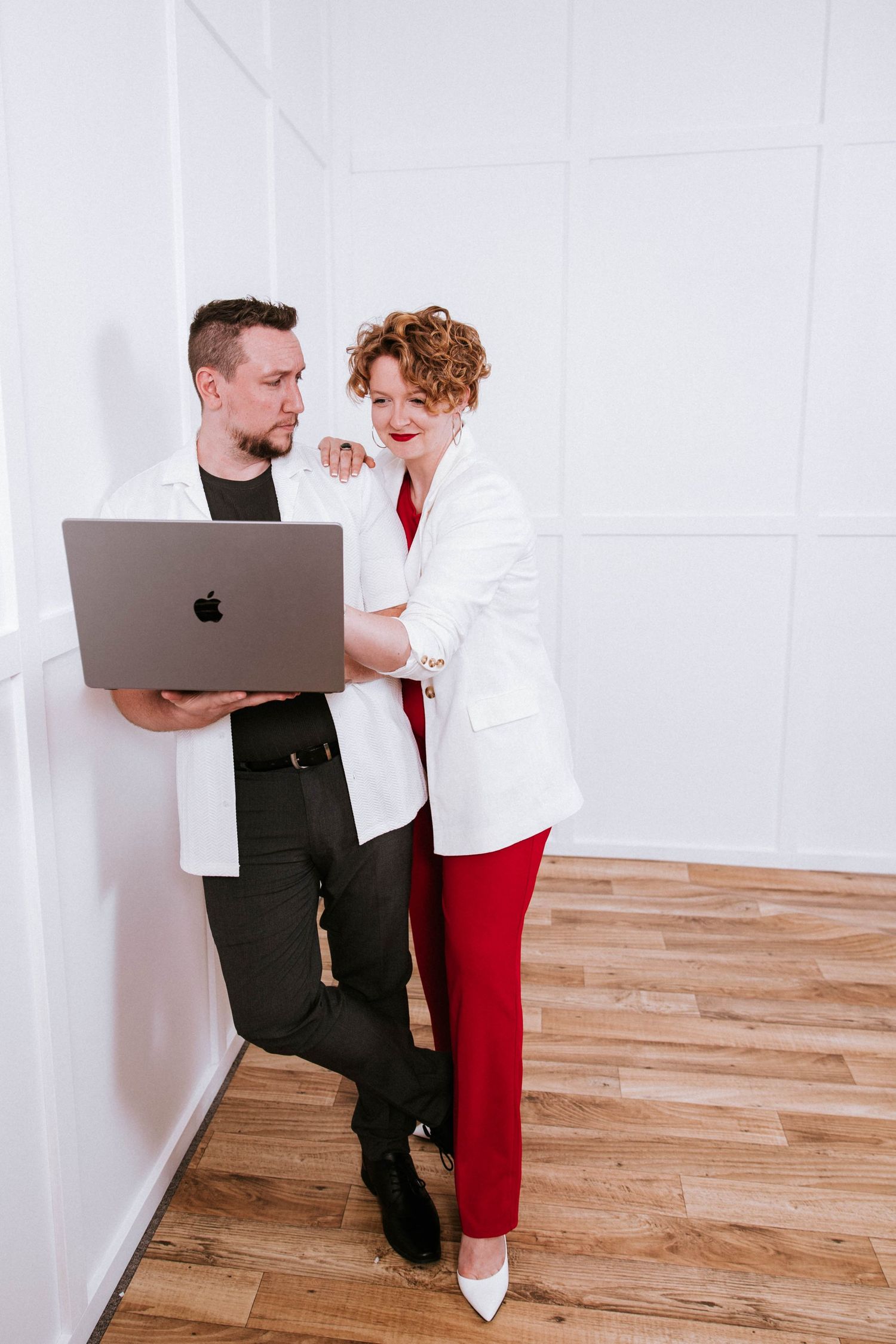 Man and woman in blazers reviewing work on a MacBook laptop while standing against a white wall.