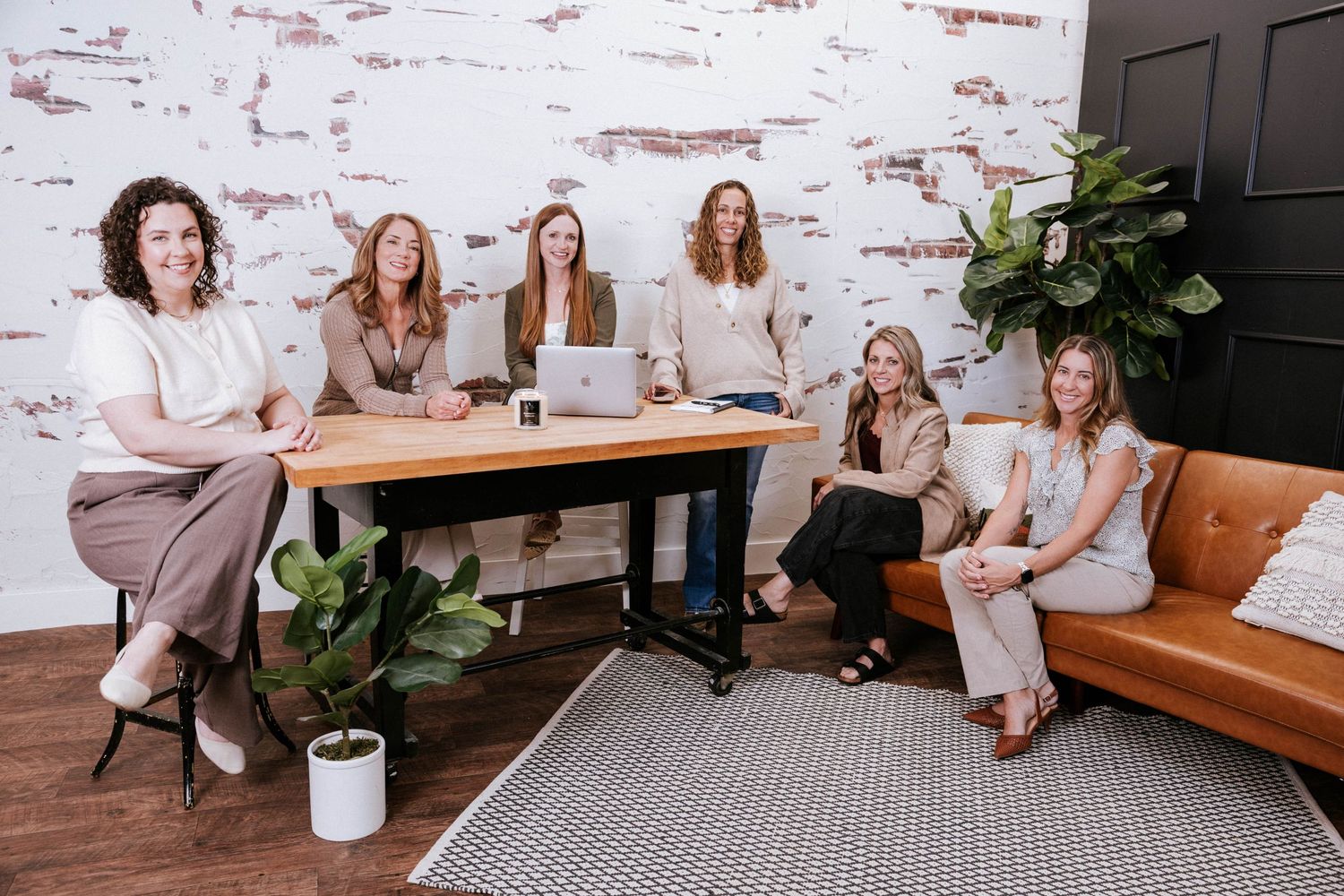 Six women pose in a modern office with brick walls, a wooden desk, laptops, and a leather sofa.
