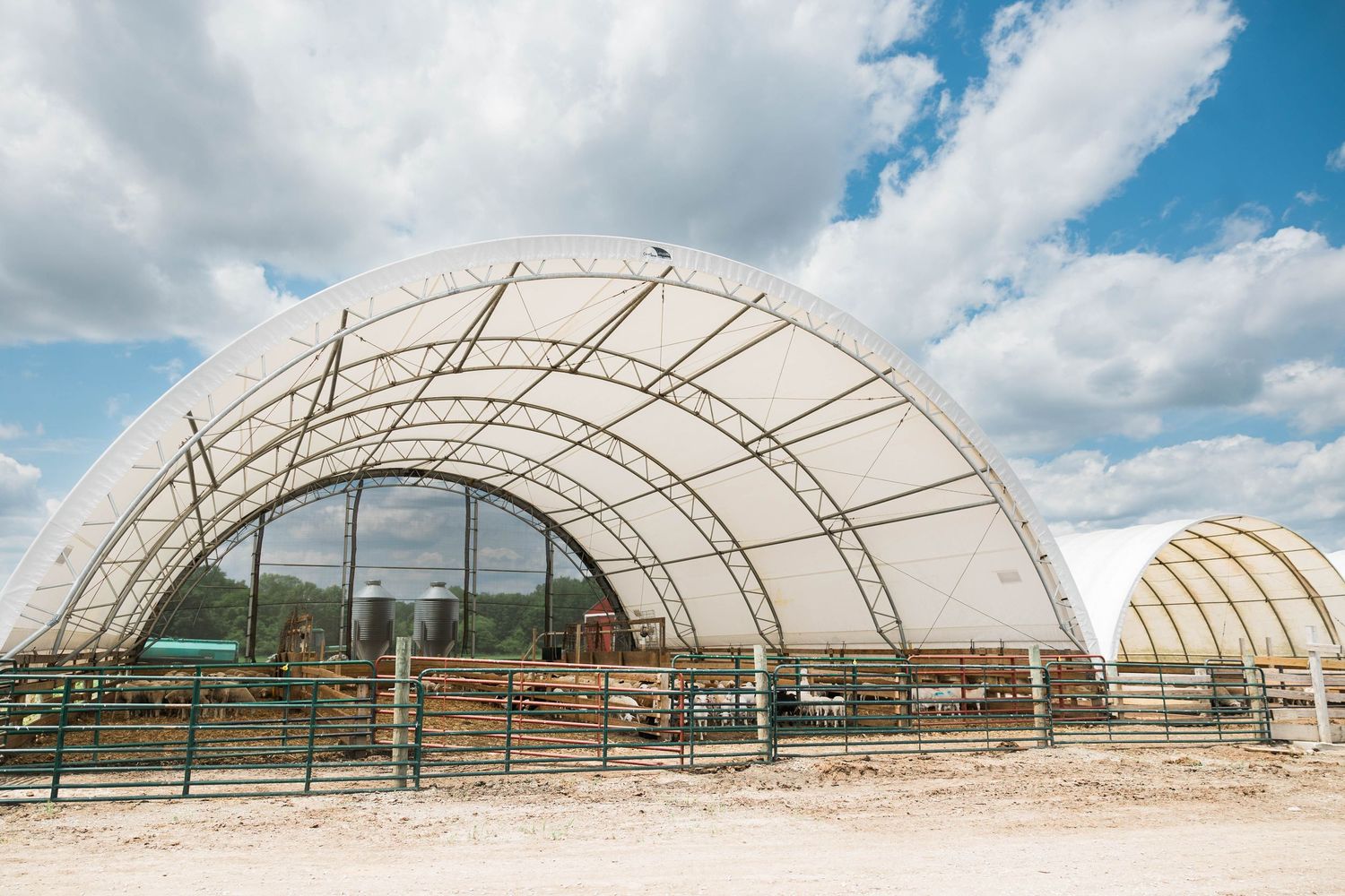 Large dome-shaped agricultural shelter with metal framework and animals in pens under cloudy blue sky.