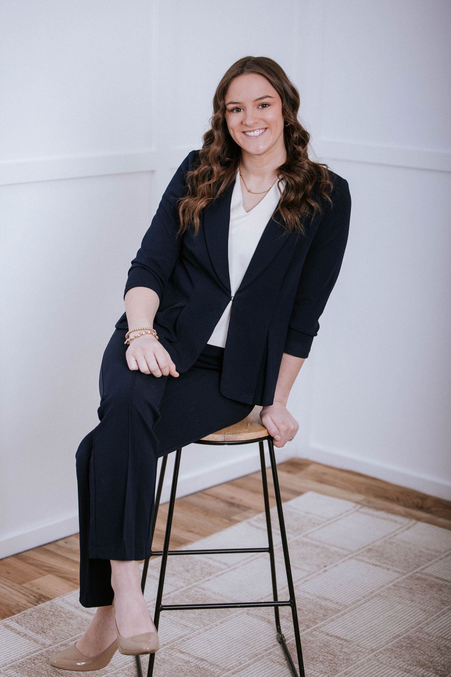 Young professional woman in navy blue suit seated on a stool, smiling confidently in a bright studio setting.